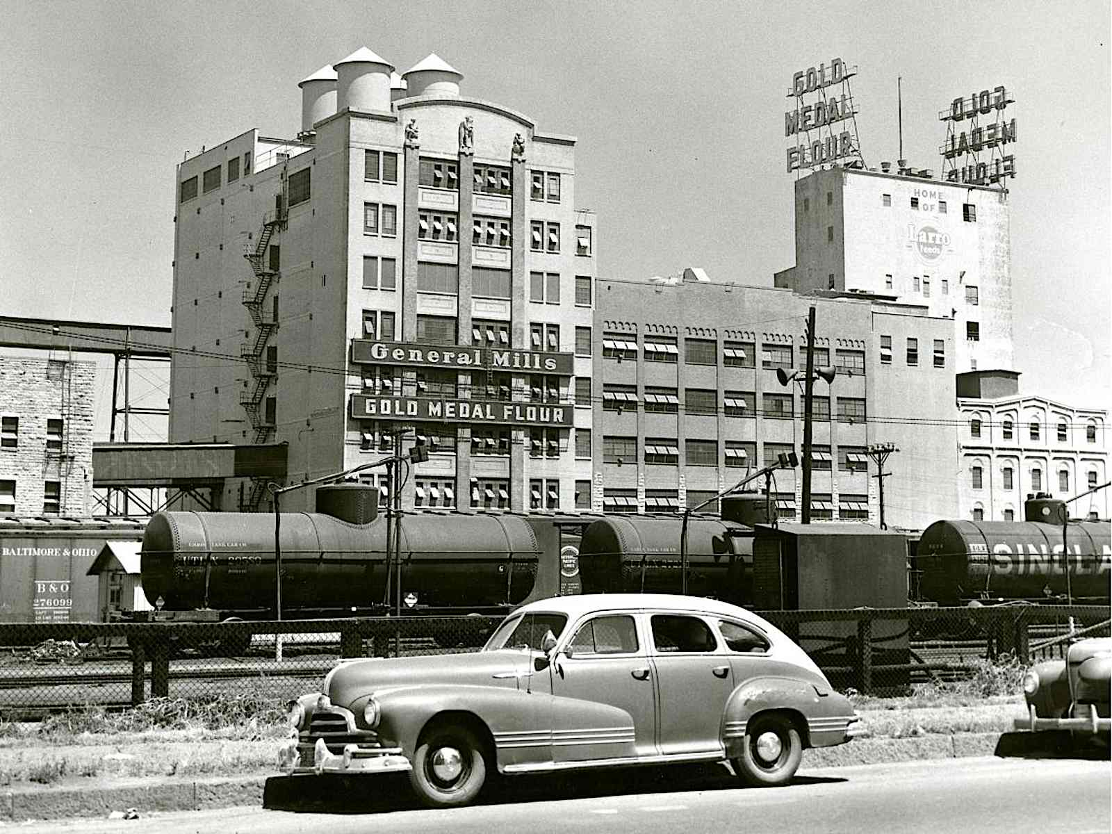A black and white photograph shows the General Mills building with "General Mills" and "Gold Medal Flour" signs prominently displayed. On the roof of a taller building behind it, large signs spell out "GOLD MEDAL FLOUR." In the foreground, a vintage car is parked on the side of a road, and behind it, several train cars, including tank cars and a boxcar are visible on tracks.